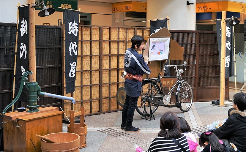 昭和３０年代の街角セットの前で紙芝居を上演する時代屋スタッフとそれを観覧するお客様たちの写真